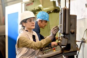 Male and female manufacture workers working on electronic machine, wearing hard hats.