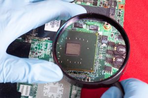 Gloved hand holding a magnifying glass and examining a printed circuit board. 