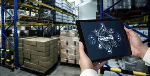Closeup of hands holding a tablet that says "Compliance" in a warehouse with shipping boxes 