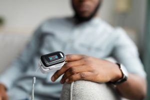 Blurred image of a man sitting on couch taking his pulse with an oximeter