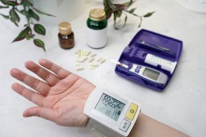 Closeup of a patient's hand, monitoring their blood pressure at home with a glucose monitor lying next to their hand.